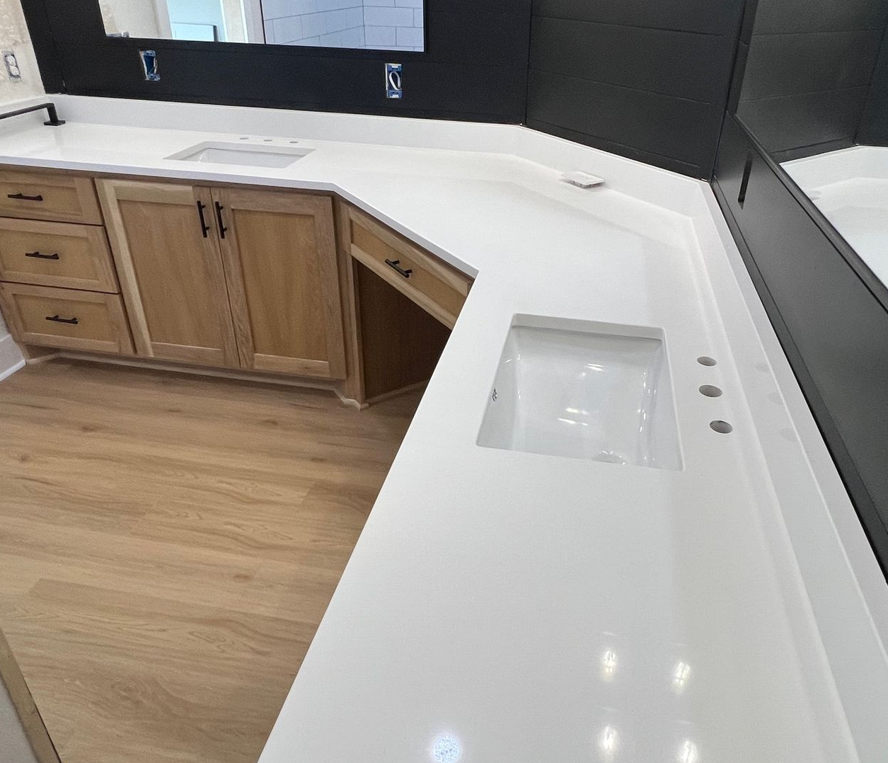 A double-sink bathroom vanity with wood cabinets, white countertops, black wall tiles, and mirrors in a bright room.