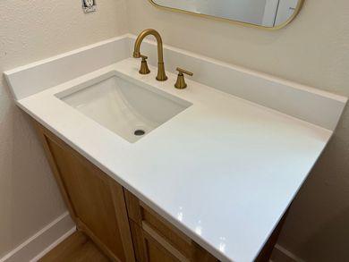 Modern bathroom vanity with white countertop, undermount sink, gold faucet, and wood cabinet below.