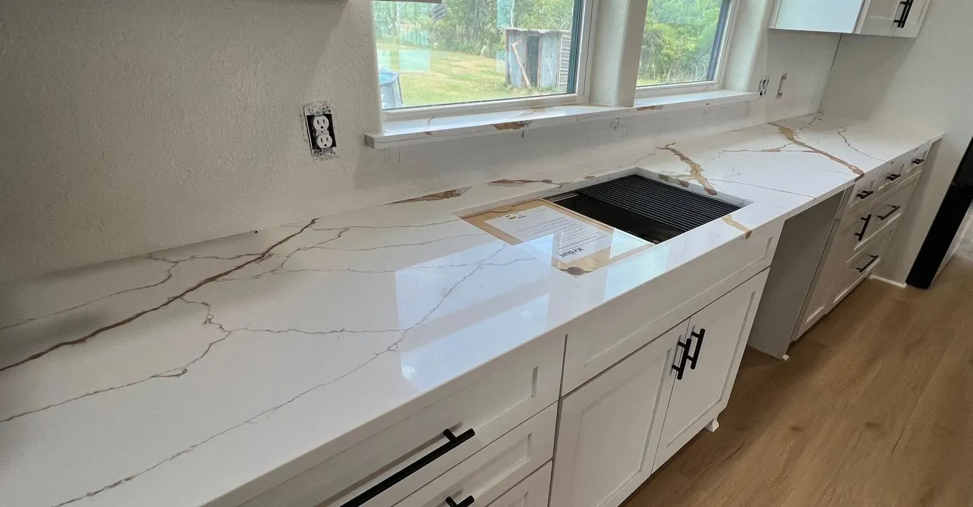 White kitchen countertop with gold veining, sink, and cabinets with black hardware.