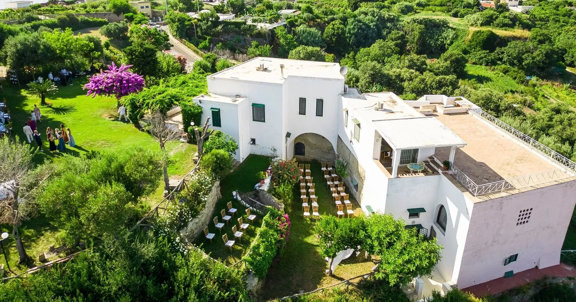 Edificio bianco su una collina circondato dal verde, con posti a sedere all'aperto predisposti per un evento.
