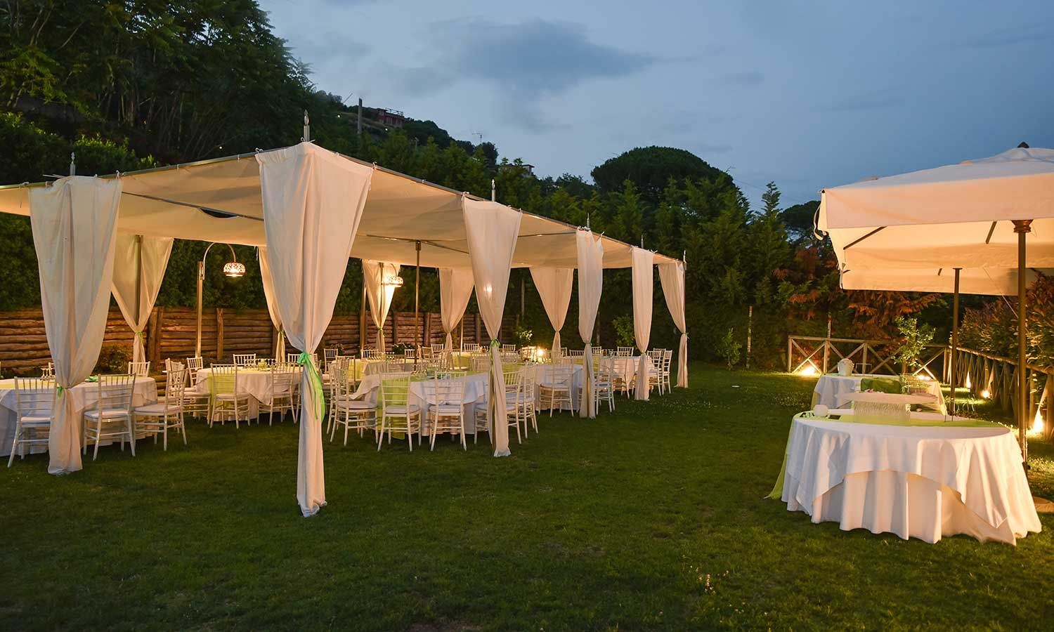 Ricevimento di nozze all'aperto con tende bianche, tavoli e decorazioni su un prato verde al tramonto.