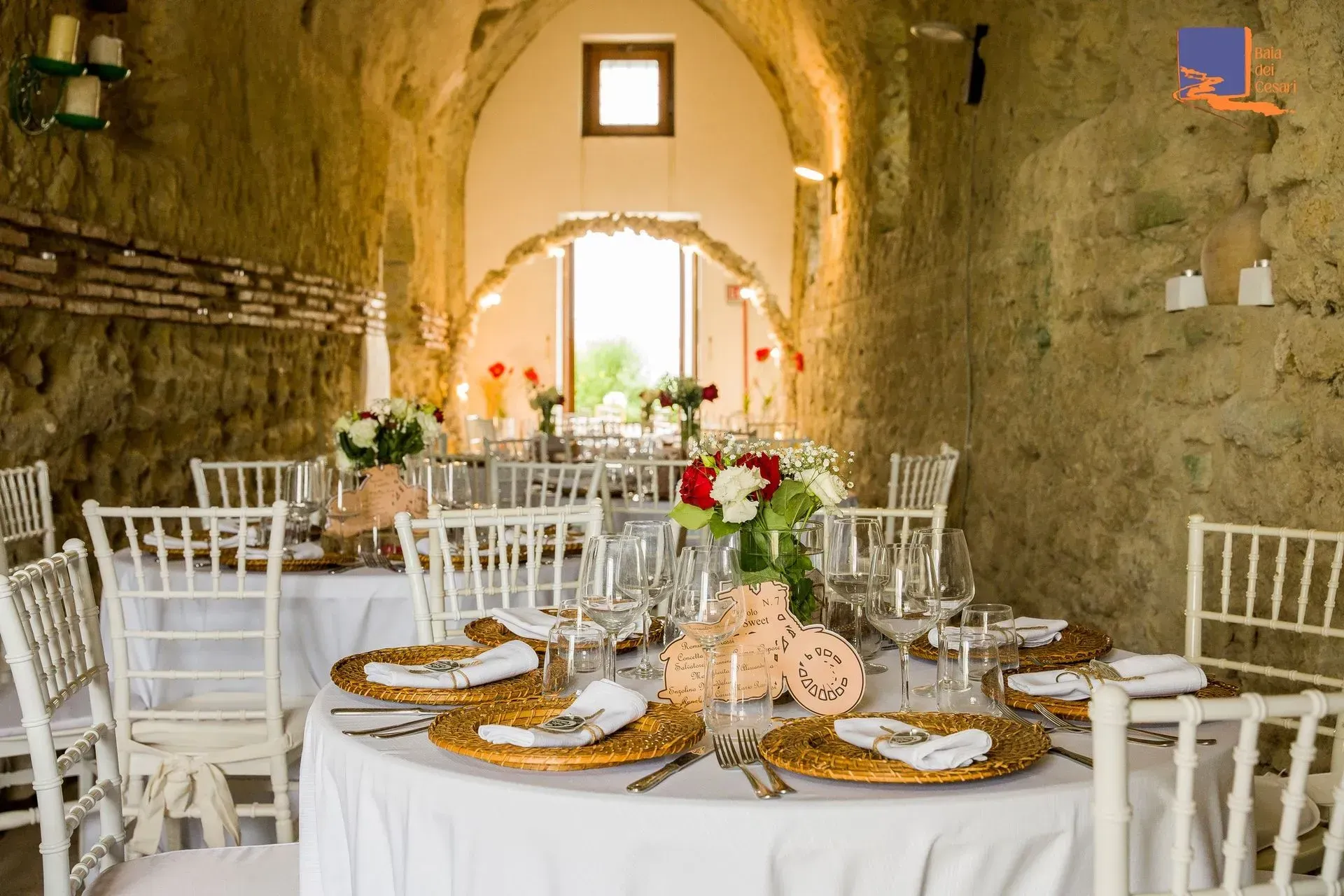 Ricevimento di nozze con tavoli rotondi, sedie bianche e centrotavola floreali in un rustico edificio in pietra.