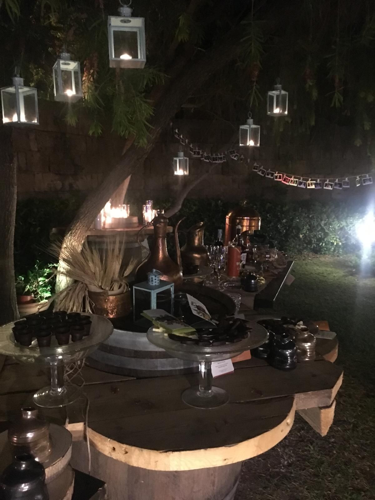Buffet di dolci all'aperto di sera, con lanterne appese a un albero. Tavoli di legno espongono cibo e bevande.