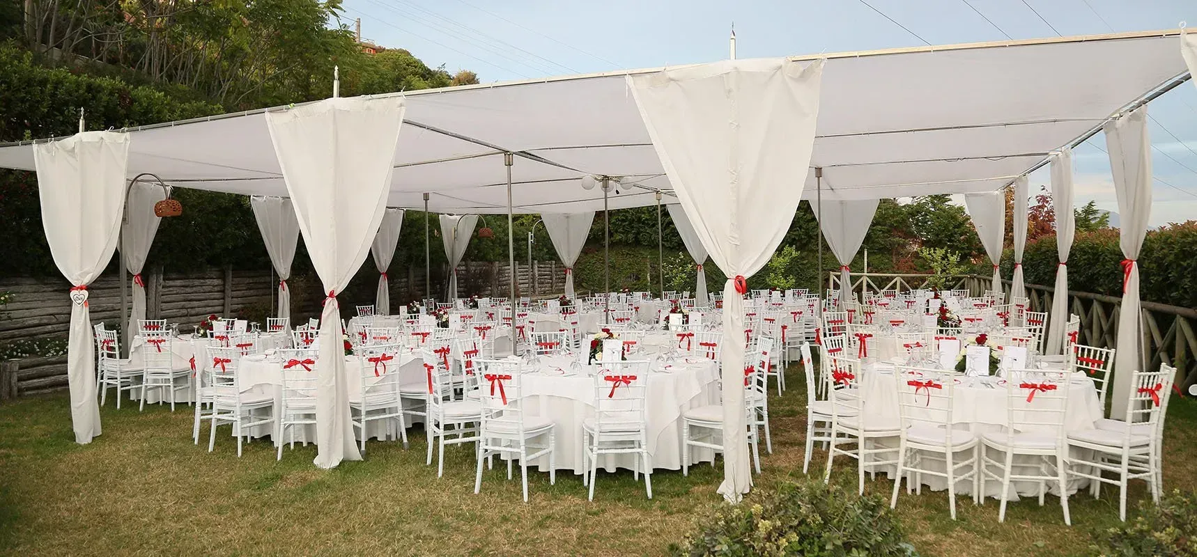 Area reception esterna con tavoli, sedie e baldacchino bianchi, decorata con dettagli rossi.