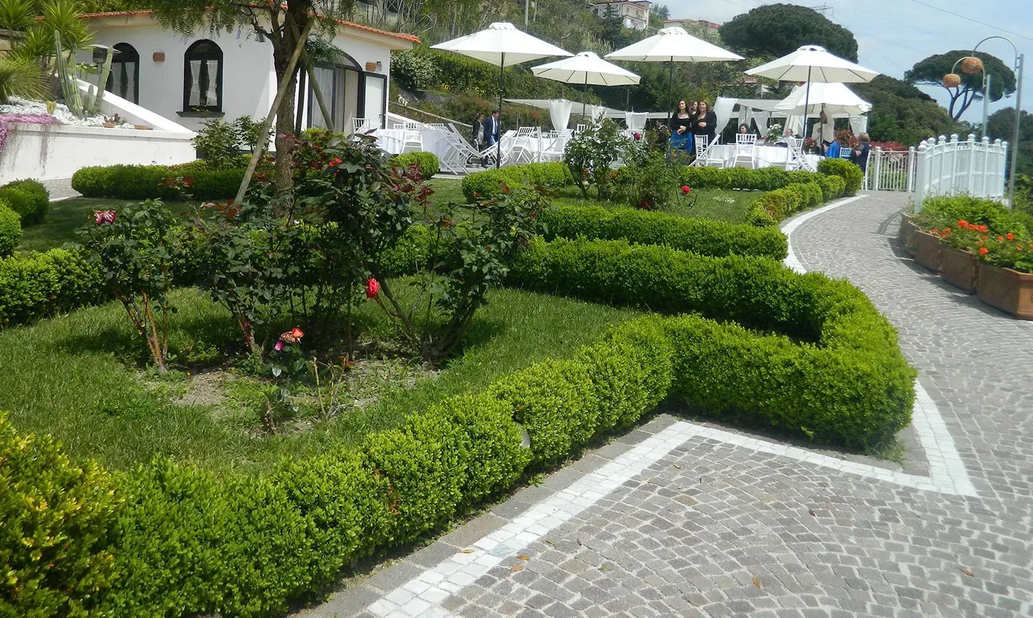 Giardino curato con siepi verdi che costeggiano un sentiero in pietra, ristorante con ombrelloni bianchi sullo sfondo.