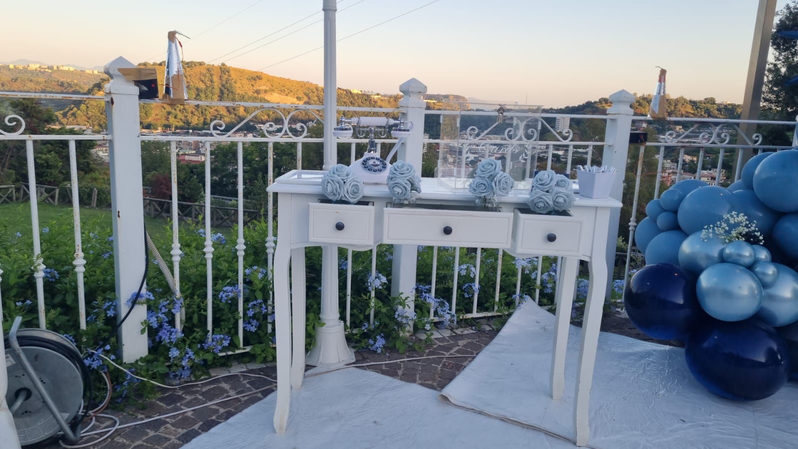 Tavolo bianco decorato con fiori blu e palloncini, all'aperto con vista panoramica.