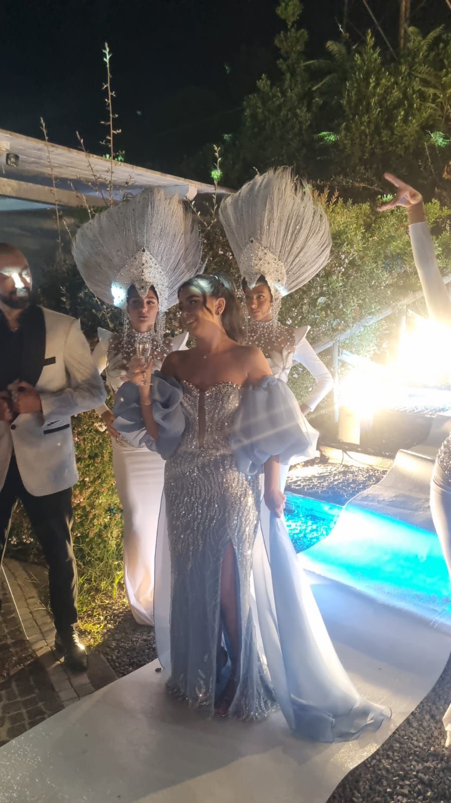 Una donna con un abito blu scintillante, affiancata da artisti vestiti di bianco, in piedi all'aperto vicino a una piscina.
