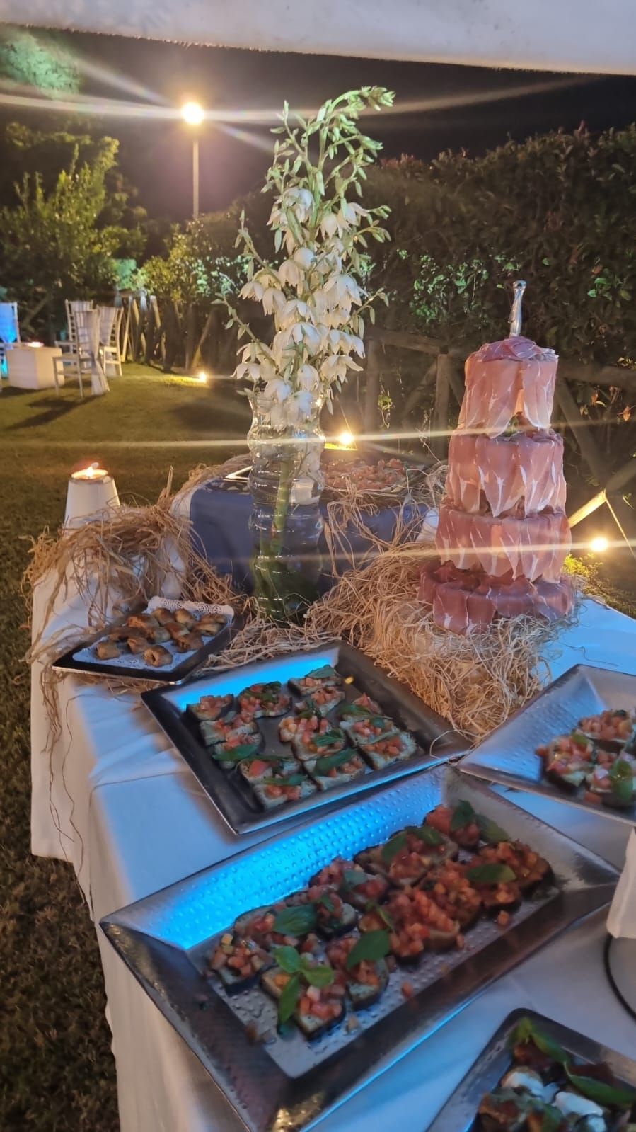 Tavolo da buffet all'aperto con vassoi di cibo, torta a piani, composizioni floreali e balle di fieno.