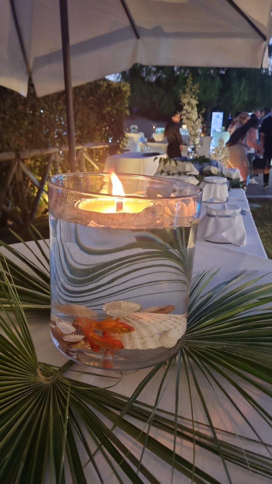 Pesci rossi in un vaso di vetro con una candela accesa, su un tavolo. Foglie di palma in primo piano. Ambientazione esterna.