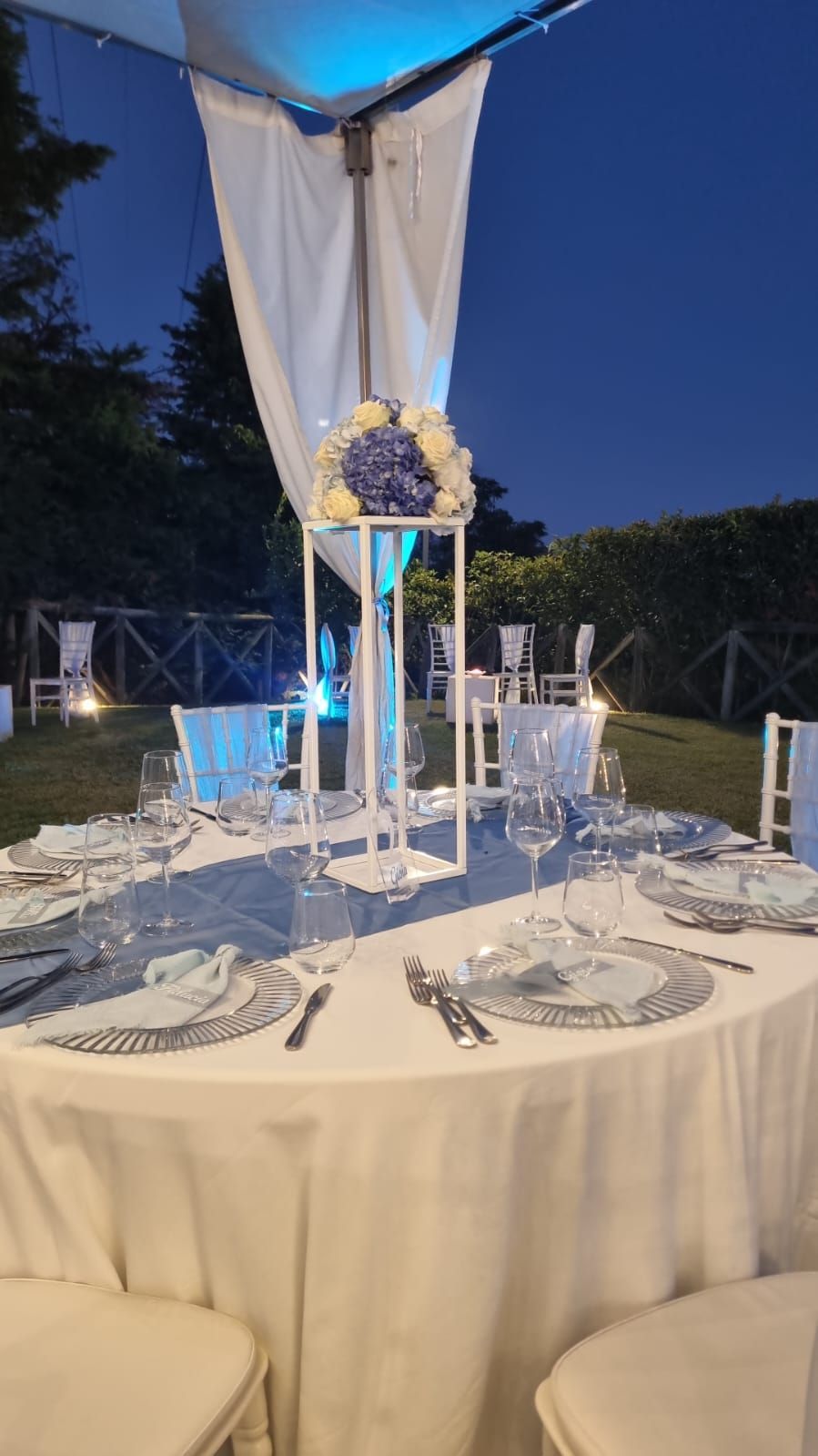 Elegante apparecchiatura per un tavolo all'aperto con centrotavola floreale sotto un baldacchino al tramonto.