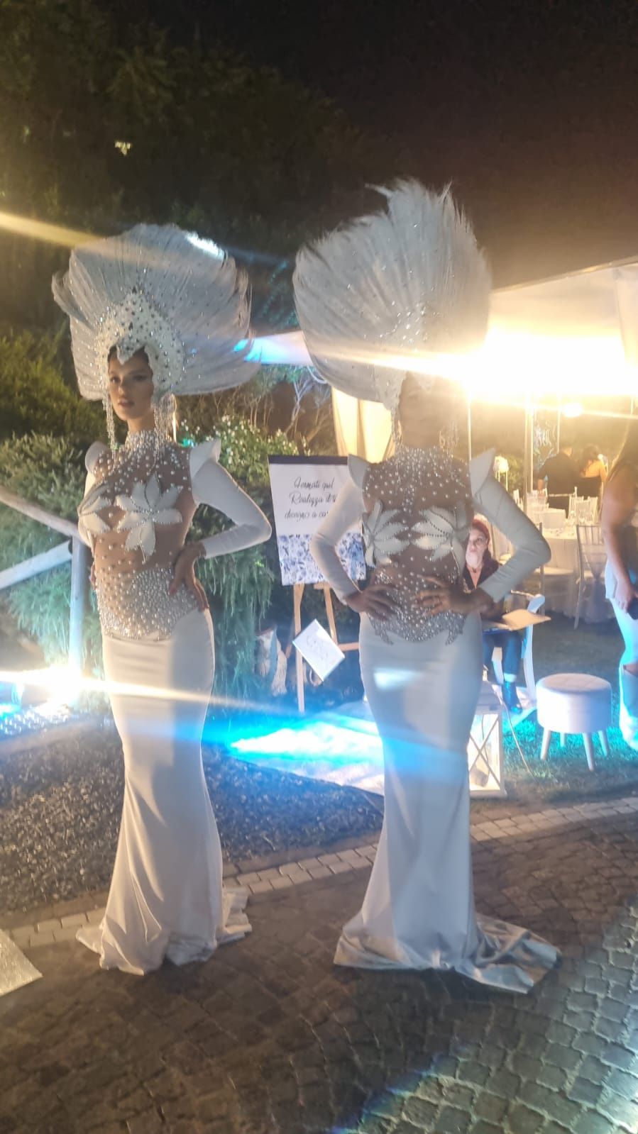 Due figure in abiti bianchi e copricapi posano all'aperto, illuminate da luci blu.