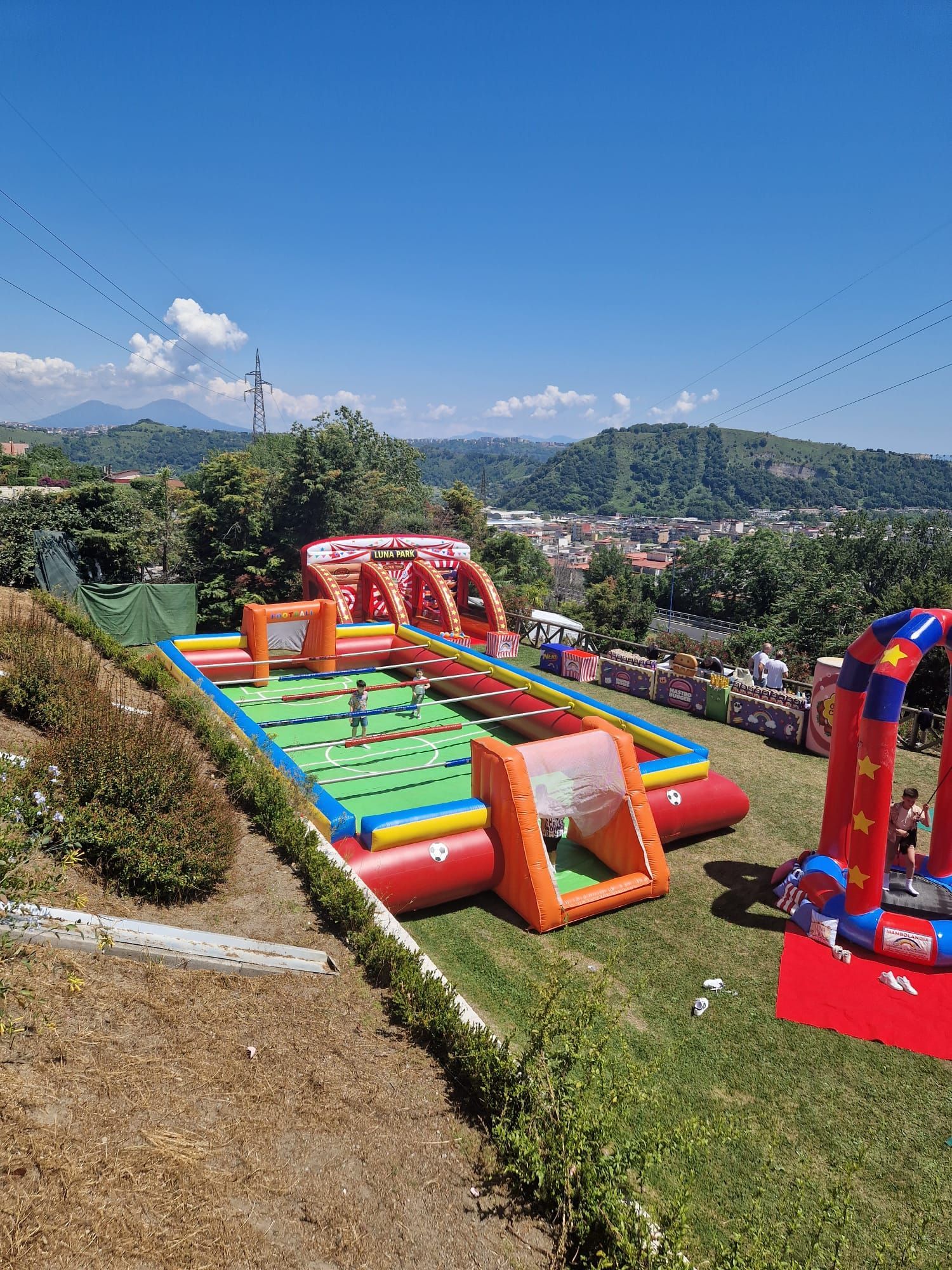Una partita di calcio gonfiabile su una collina con bambini che giocano. Colori vivaci, cielo azzurro e una città lontana.