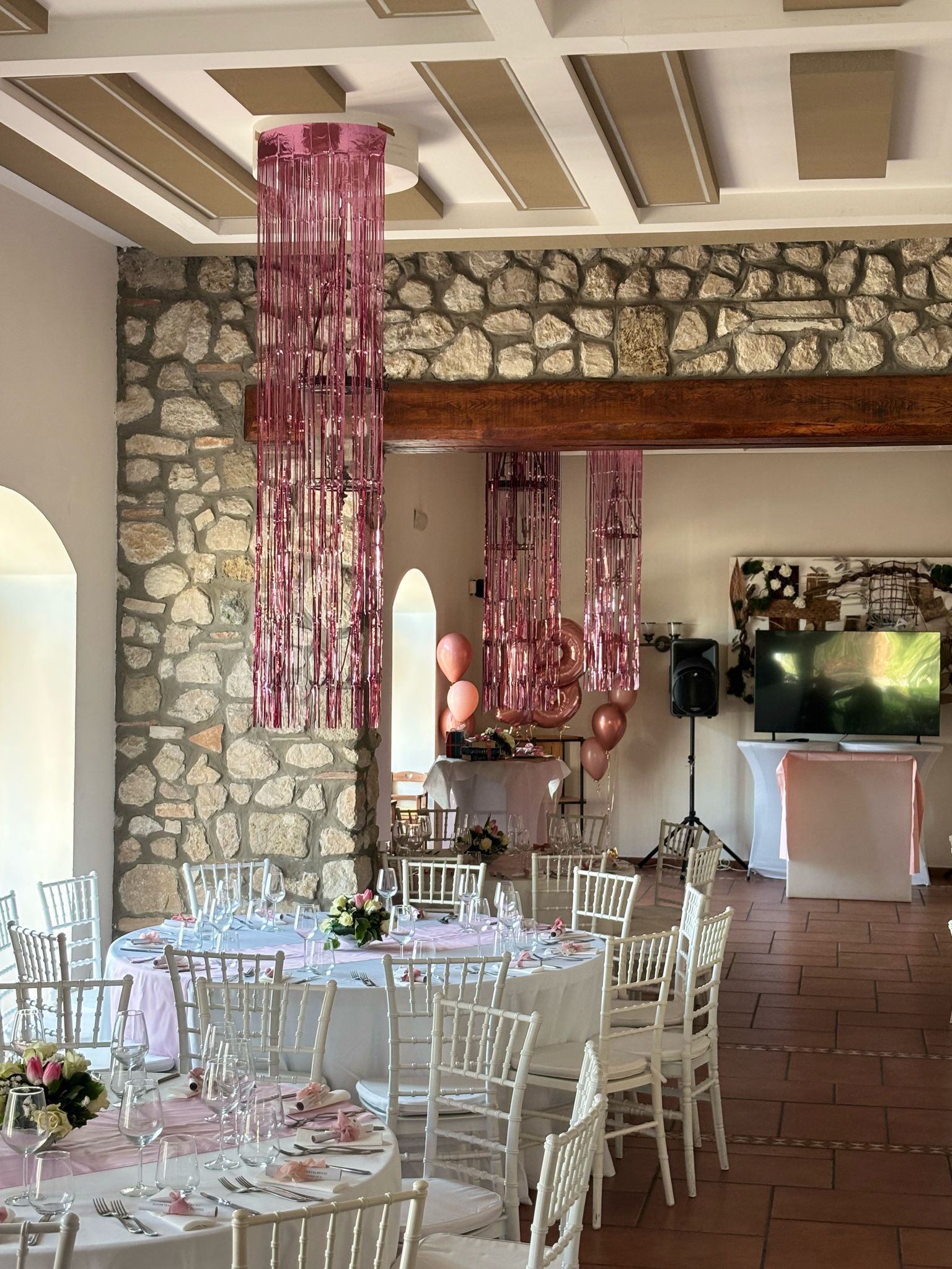 Sala da pranzo decorata con tavoli apparecchiati per una festa, con festoni rosa, palloncini e pareti in pietra.