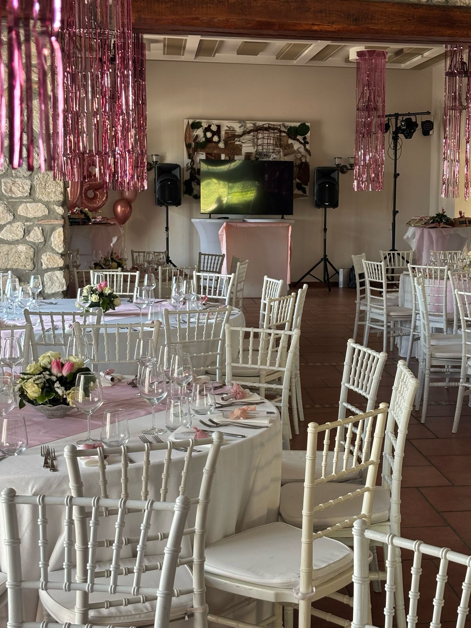Sala allestita per un evento con tavoli bianchi, sedie, decorazioni rosa e una TV.
