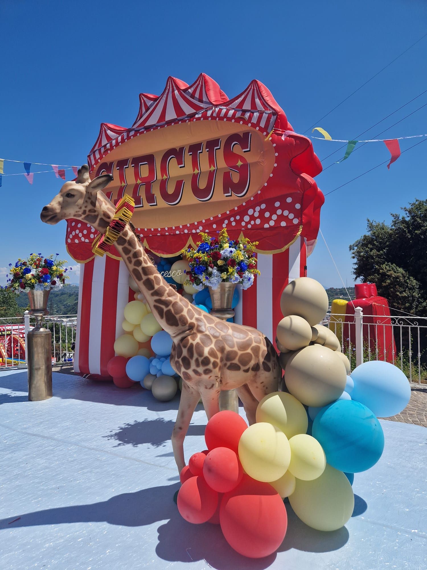 Giraffa con palloncini davanti al cartello di un circo; sfondo cielo azzurro.