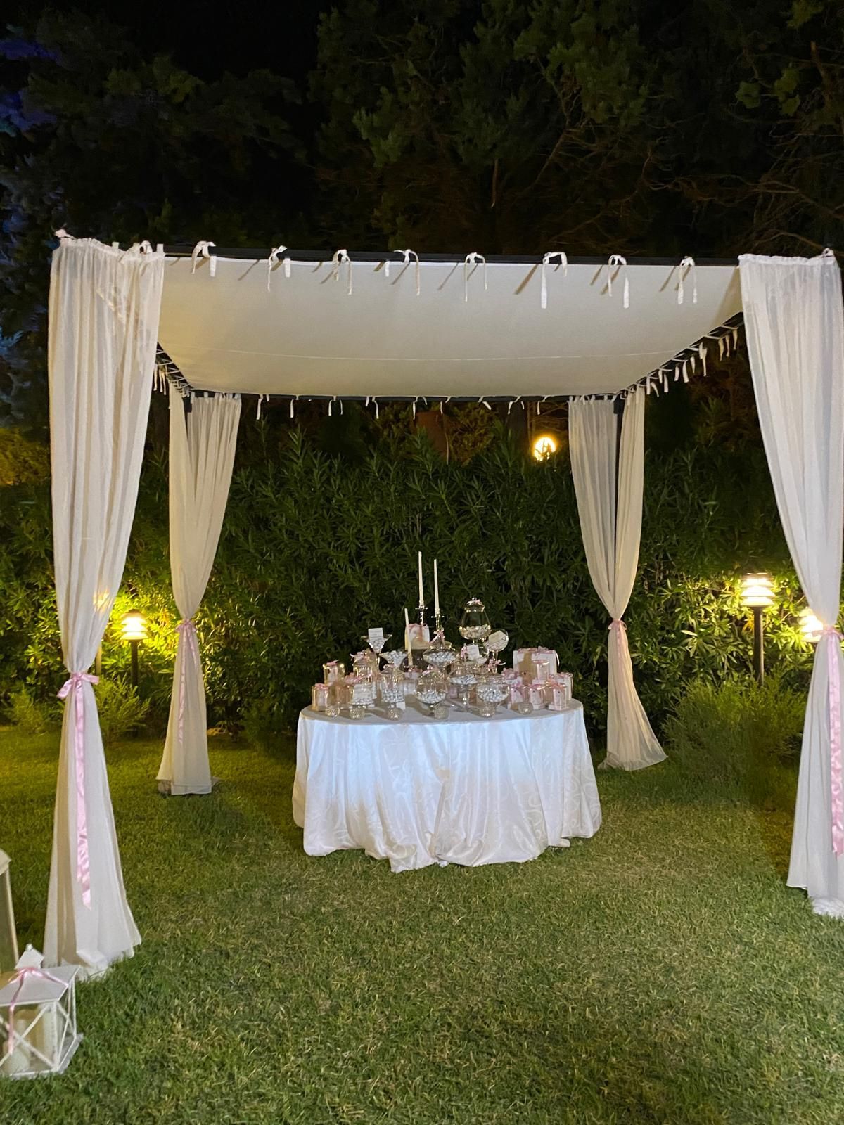 Un baldacchino bianco drappeggiato copre un tavolo apparecchiato con dolcetti, in un giardino di notte. Nastri rosa impreziosiscono le tende.
