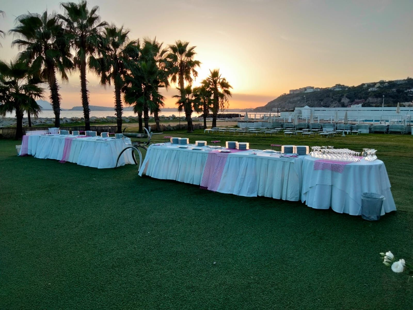 Tavoli apparecchiati per un evento all'aperto su un'area erbosa, con palme e un tramonto sullo sfondo.