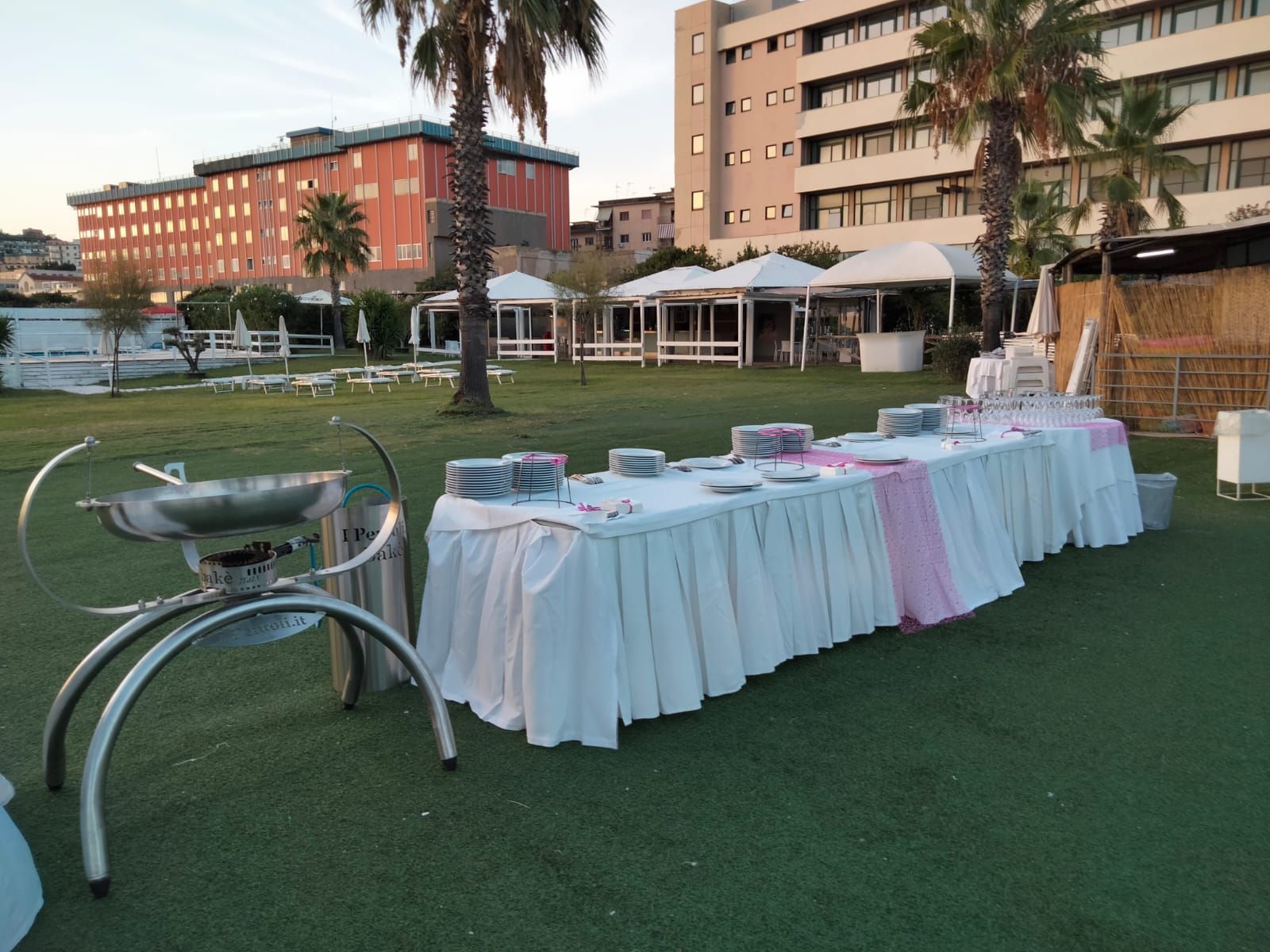 Tavolo da buffet apparecchiato per un evento all'aperto, con una tovaglia bianca e dettagli rosa, di fronte a un edificio.