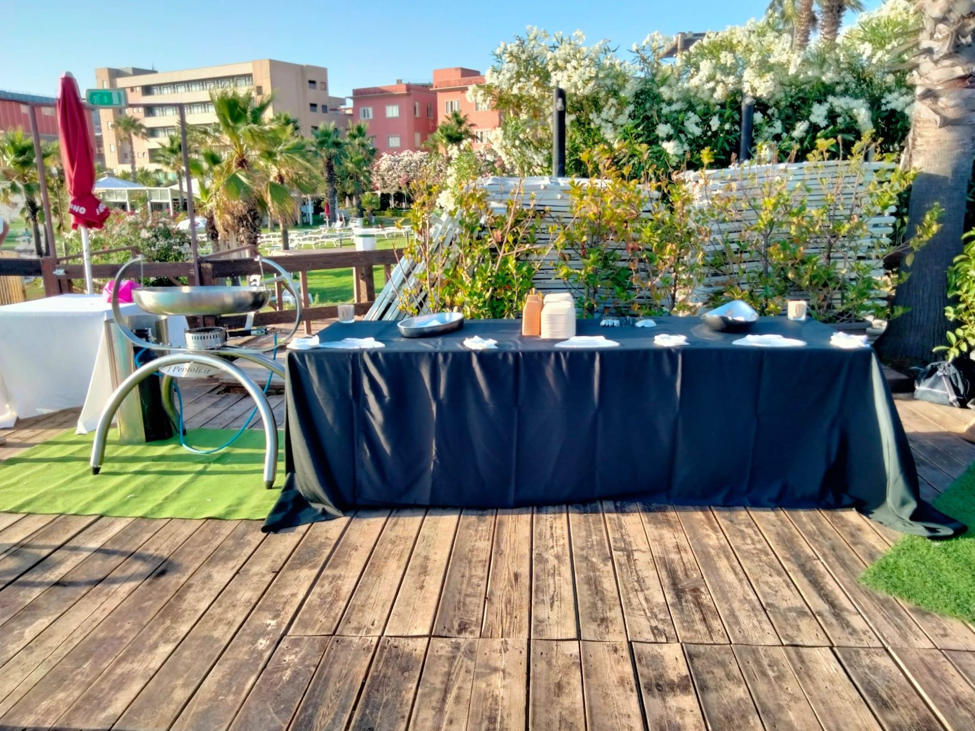 Tavoli da pranzo all'aperto su una terrazza in legno con tovaglie bianche e nere, circondati da piante.