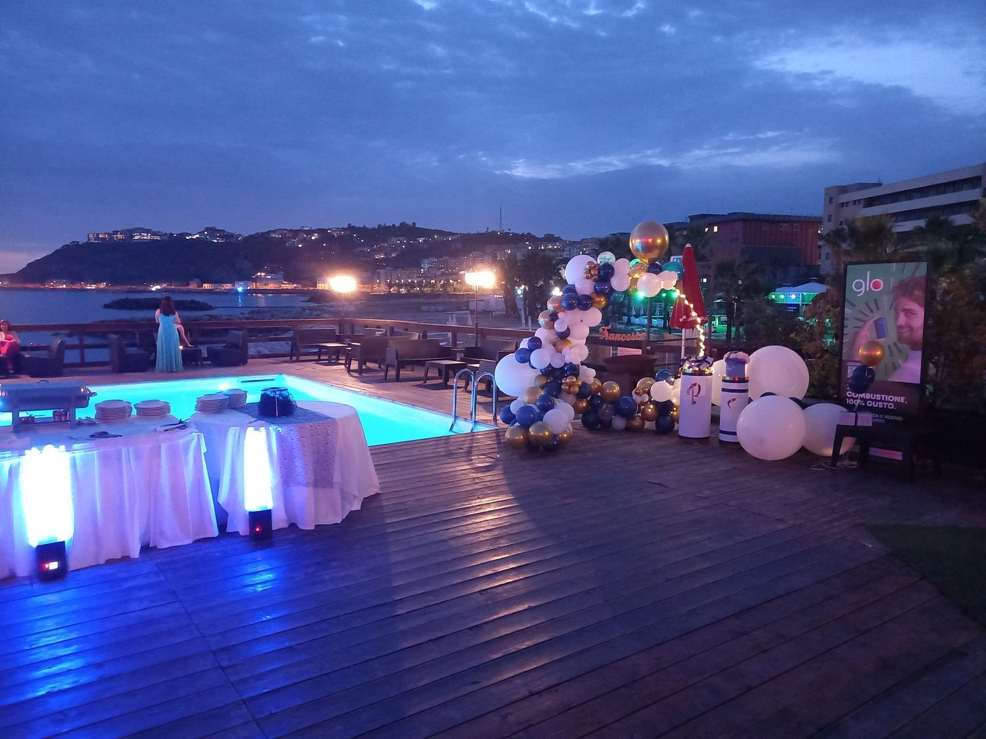 Festa all'aperto al tramonto con piscina, tavoli, palloncini e vista sulla costa.