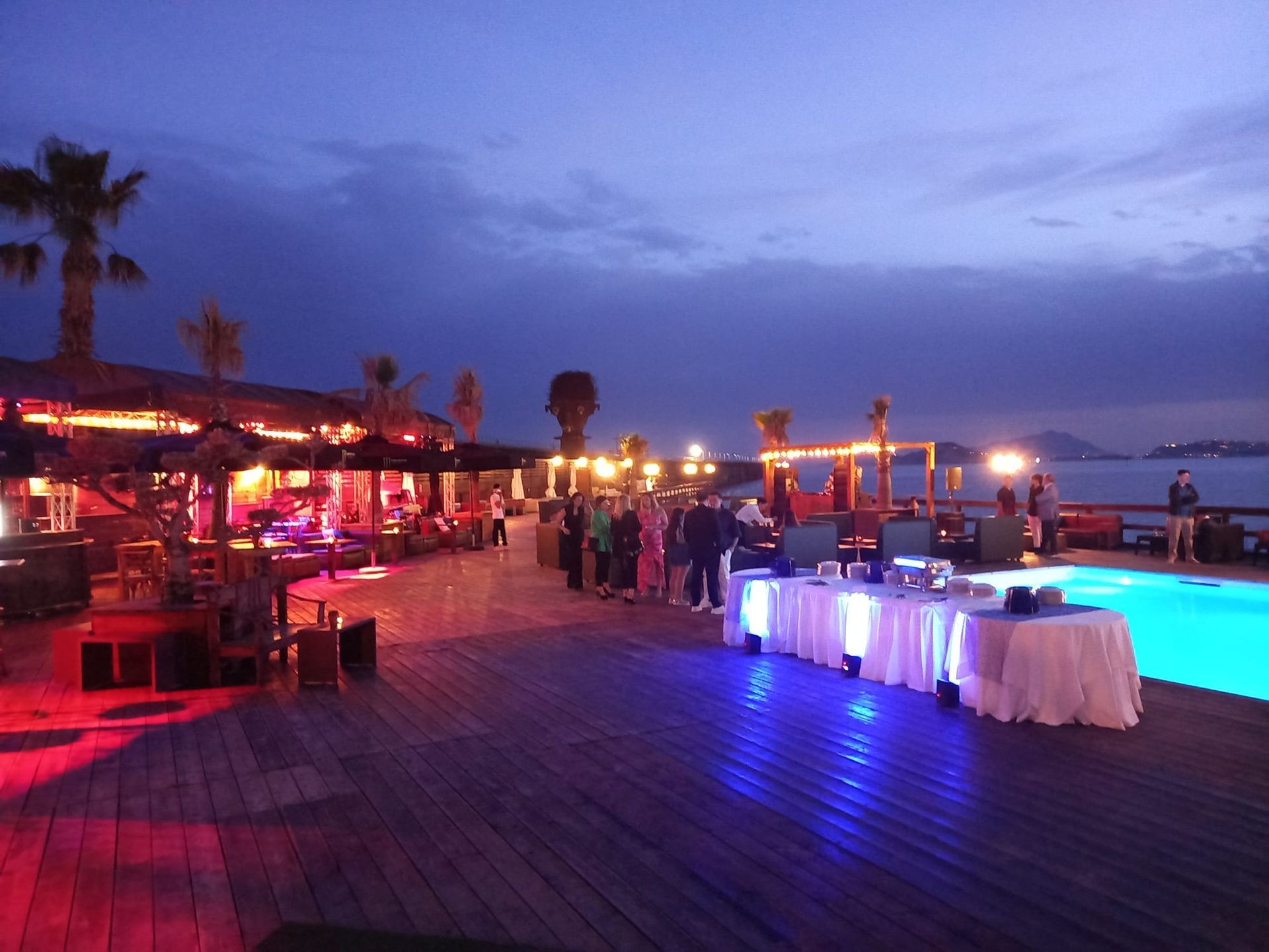 Evento notturno all'aperto con tavoli illuminati vicino a una piscina, palme e persone.