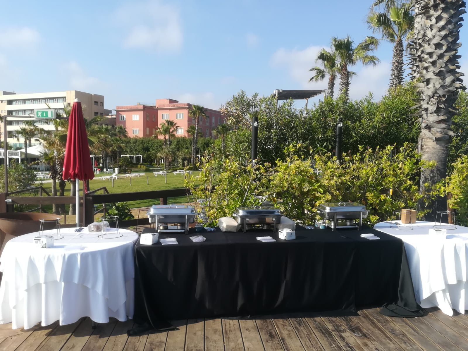 Tavolo da buffet allestito all'aperto con tovaglie bianche, palme sullo sfondo.