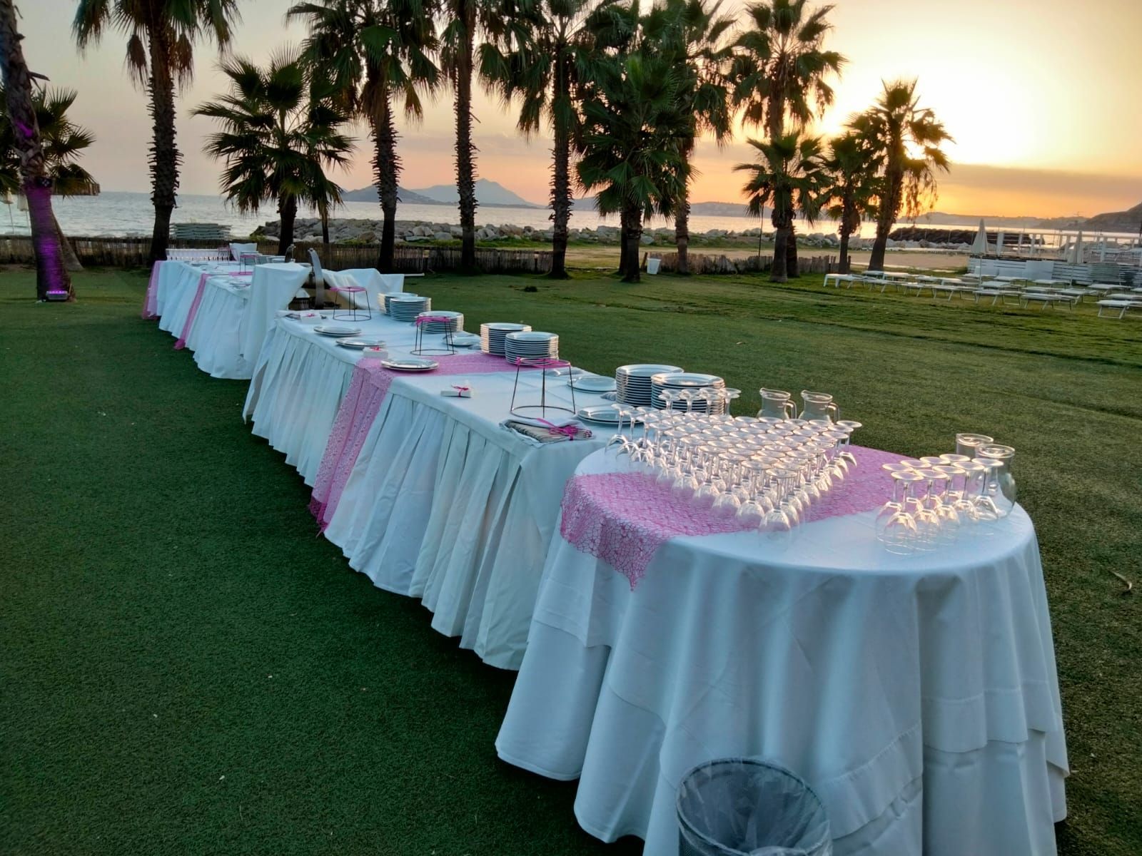 Tavoli da buffet allestiti sull'erba con sfondo di tramonto, palme e acqua.