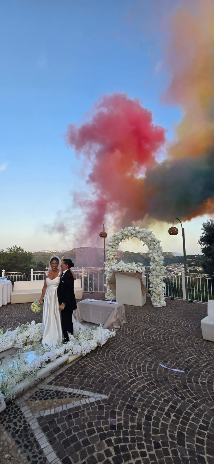 Gli sposi posano durante la cerimonia nuziale all'aperto, con fumo colorato sullo sfondo e un arco floreale.