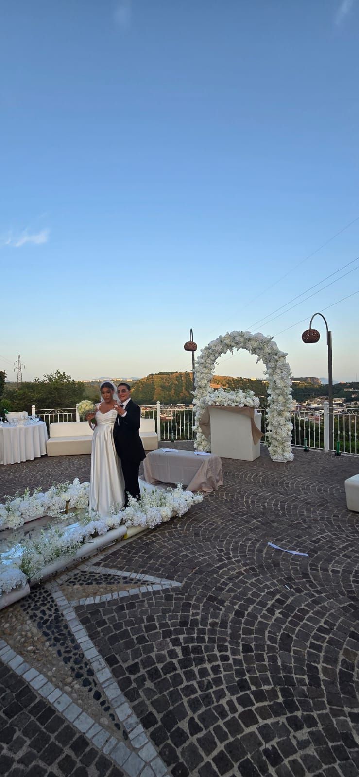 Gli sposi sono in piedi su un patio acciottolato. L'arco nuziale è decorato con fiori. Sullo sfondo, il cielo azzurro 