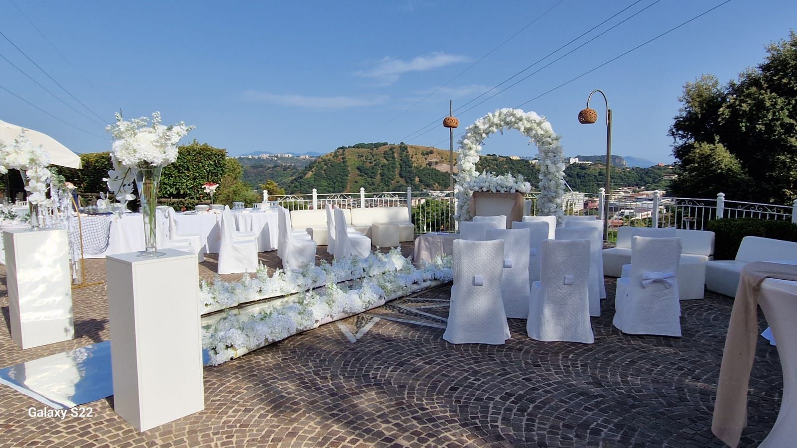 Allestimento per la cerimonia nuziale: sedie bianche, arco, composizioni floreali su una terrazza con vista su un paesaggio pittoresco.