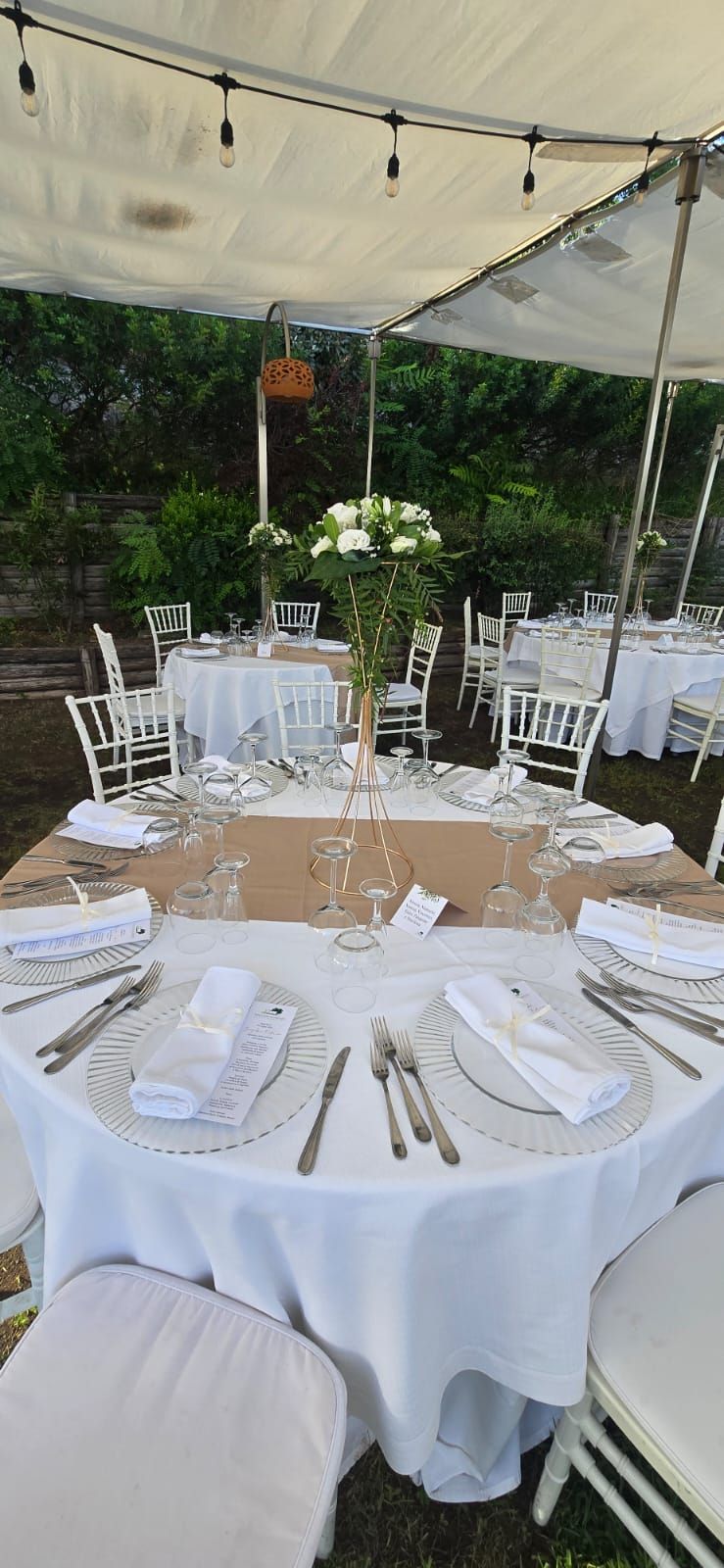 Tavoli apparecchiati per un evento all'aperto sotto una tenda, con tovaglie bianche, fiori e apparecchiature.