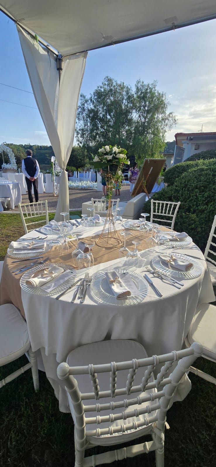 Tavolo rotondo apparecchiato per un evento formale all'aperto, coperto da un baldacchino bianco. Anche le sedie sono bianche.
