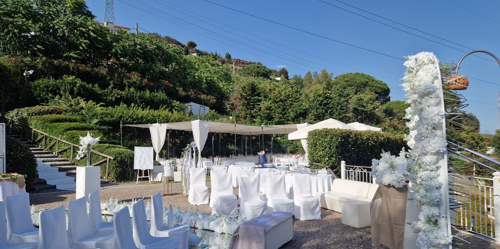 Cerimonia nuziale allestita all'aperto con sedie bianche, arco floreale e area tendata, con vista sul verde.