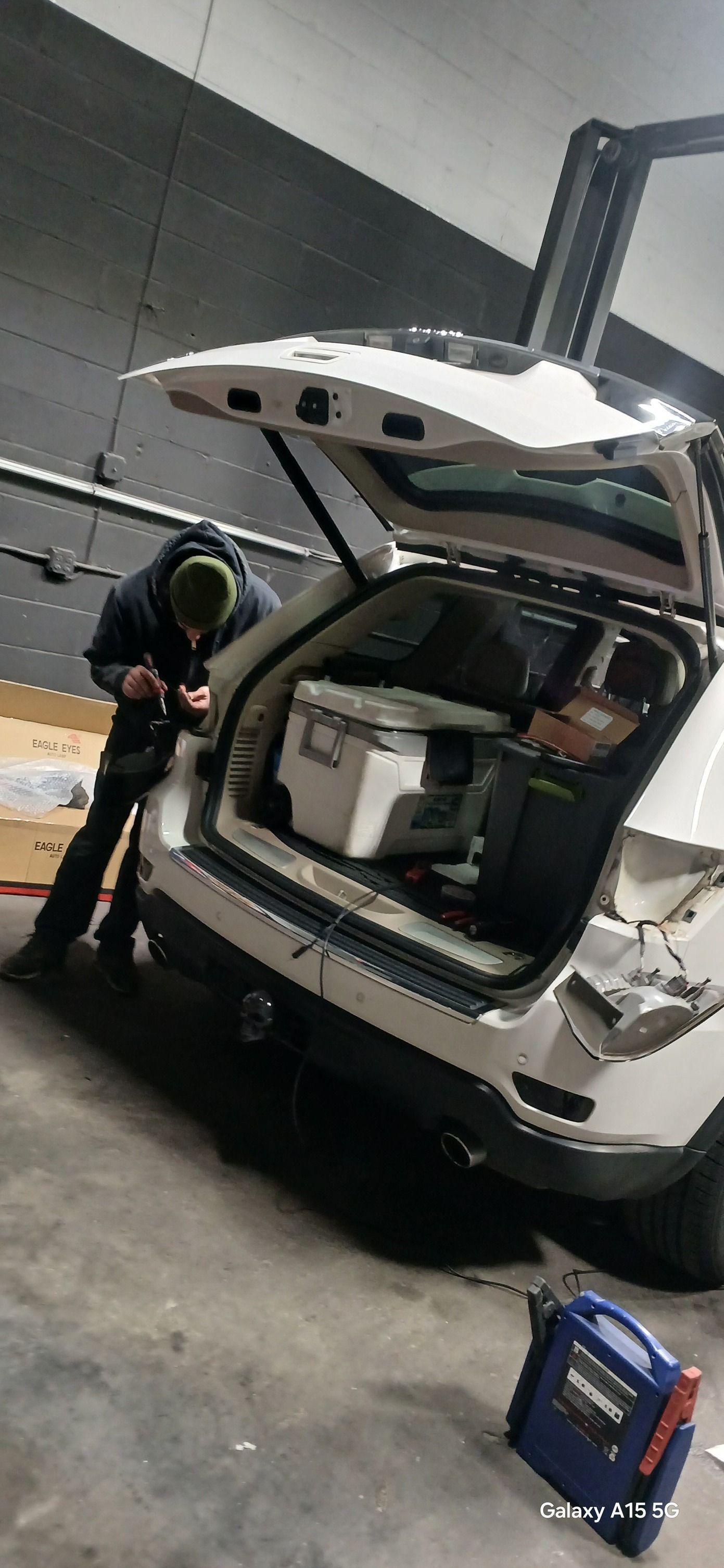 Person working on the open trunk of a white SUV with a cooler inside; a generator is on the ground.