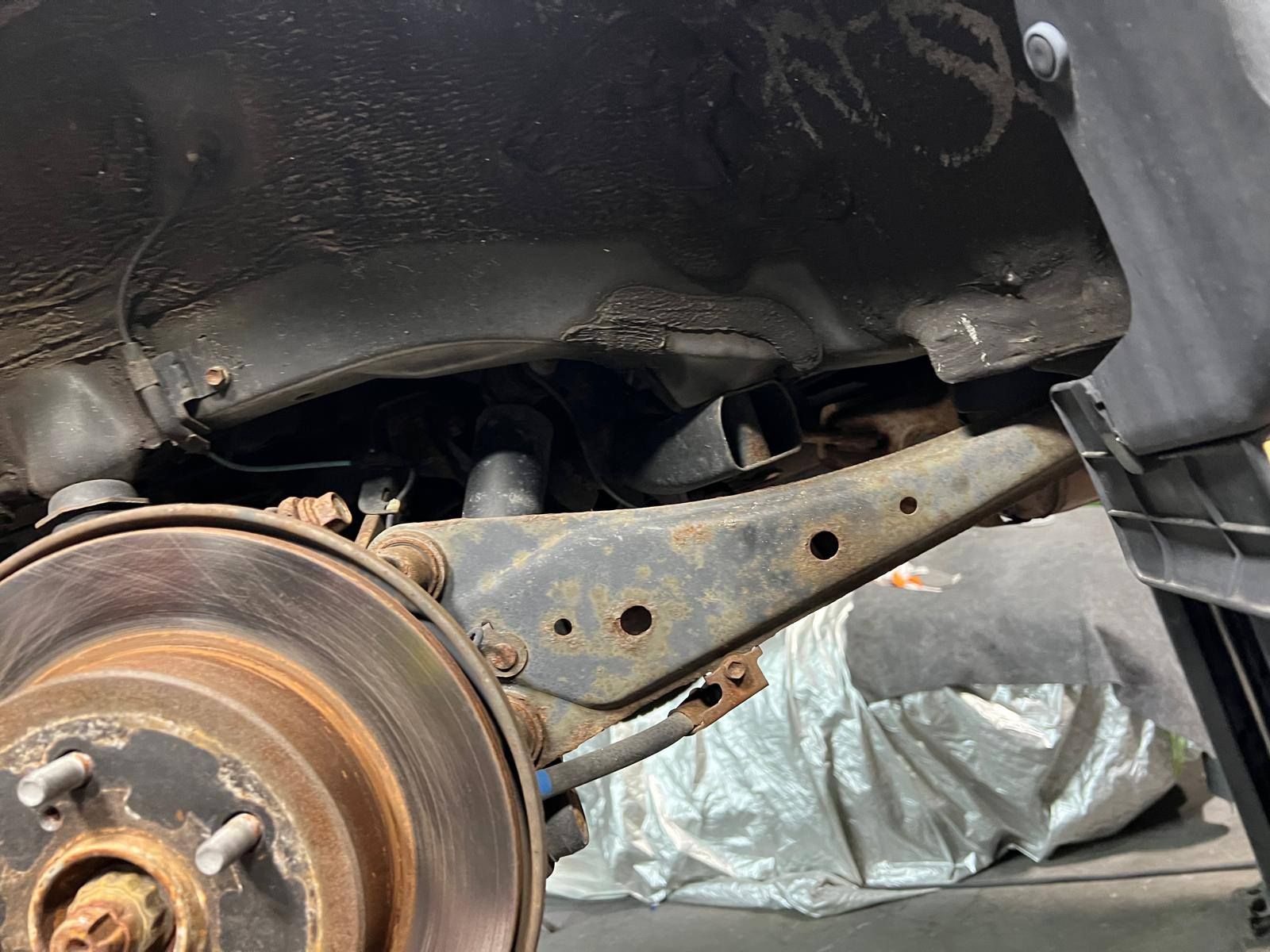 Rusty car suspension components visible from below. A wheel with a brake rotor is in the foreground.