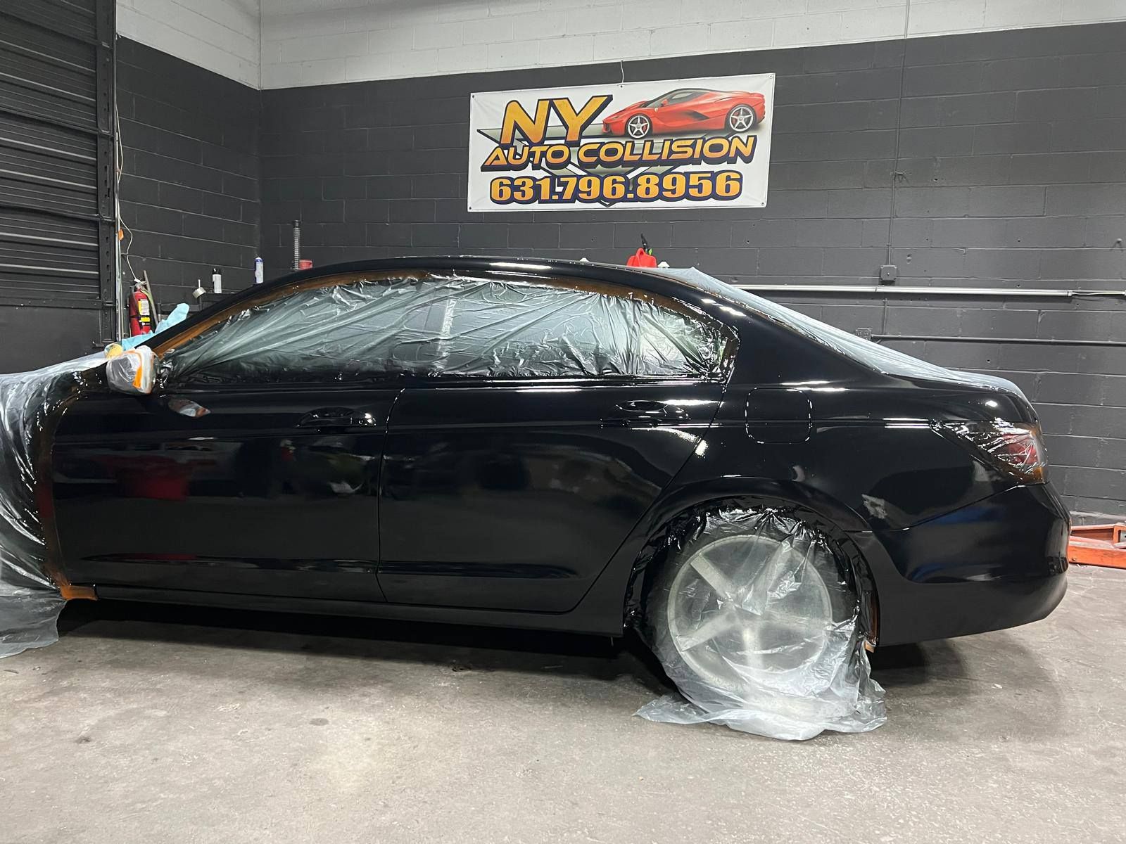 Black car covered in plastic sheeting inside a repair shop.