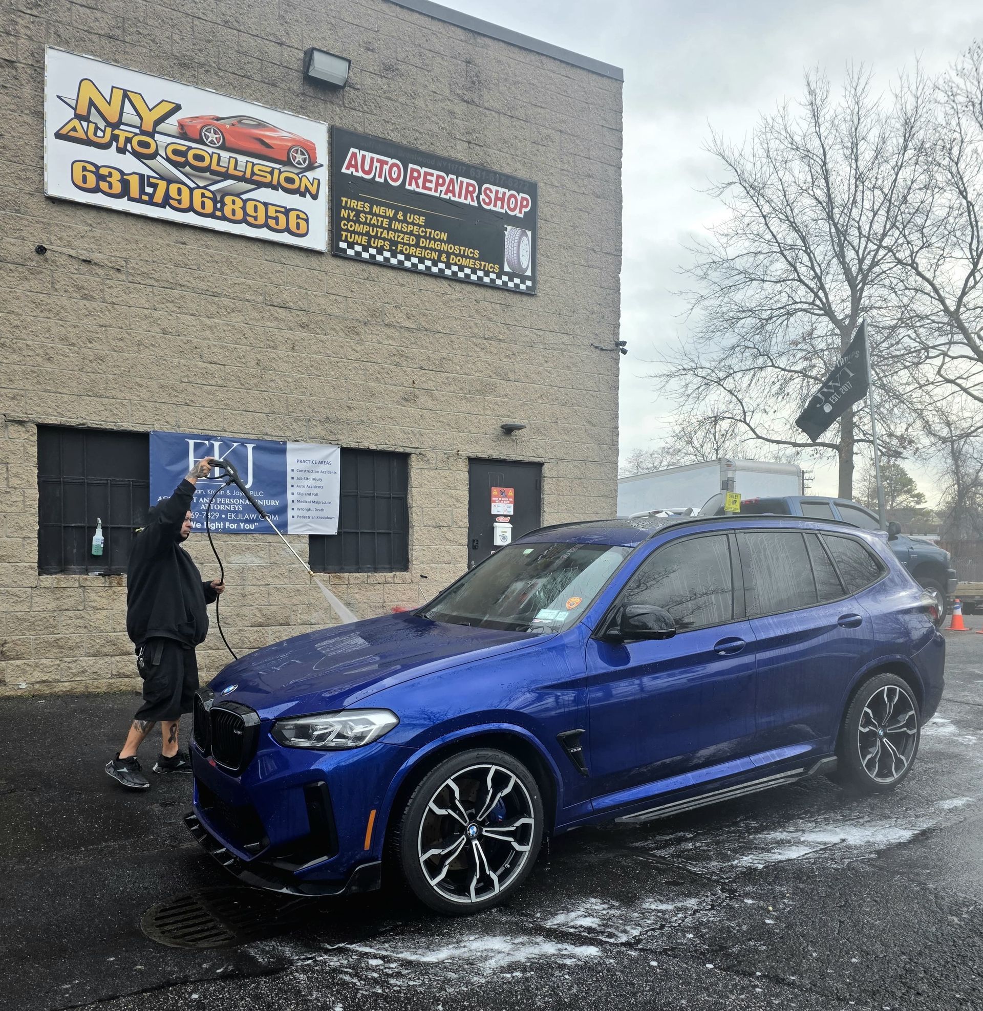 Man washing a blue BMW SUV at NY Auto Detailing, outside a brick building.