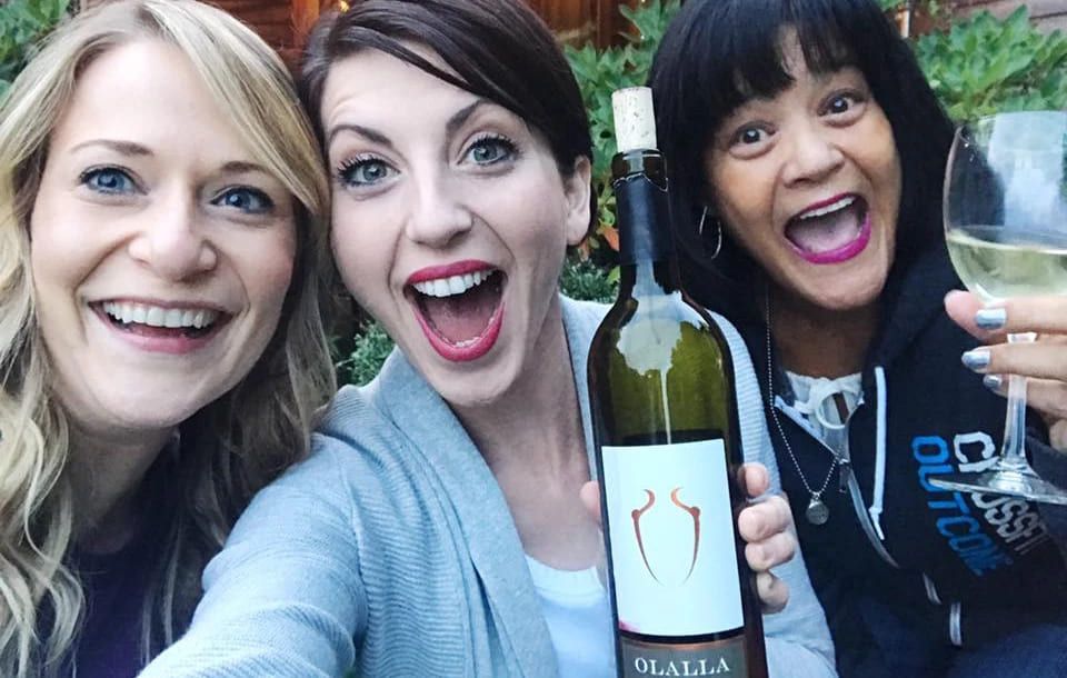 Three women are posing for a picture with one holding a bottle of wine