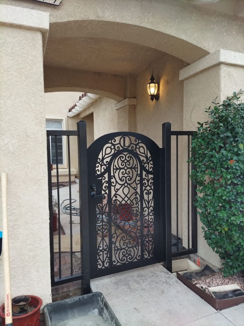 Black wrought-iron gate within a beige archway. To the right, a green bush. Cement pathway.
