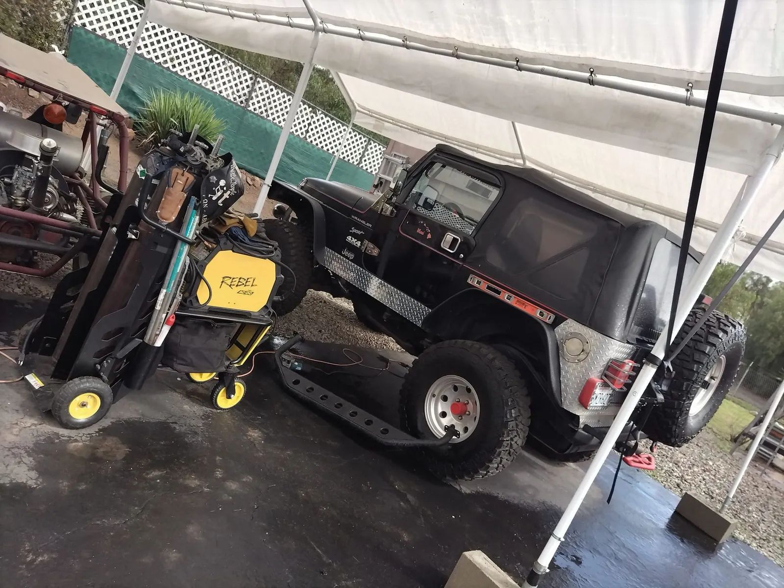 Black Jeep raised on a platform, next to a cart with equipment, under a white canopy.