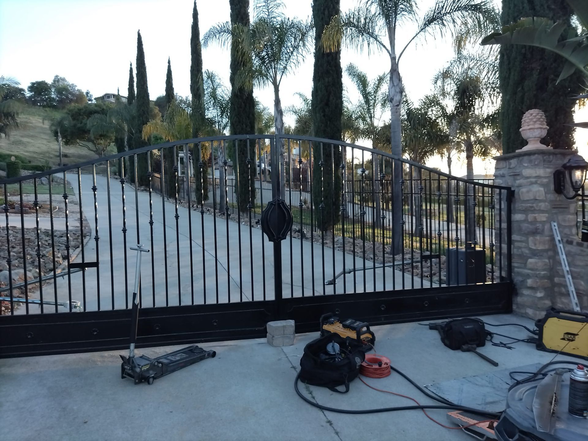 A black wrought-iron driveway gate under construction, with tools scattered on the ground in front of a stone pillar.