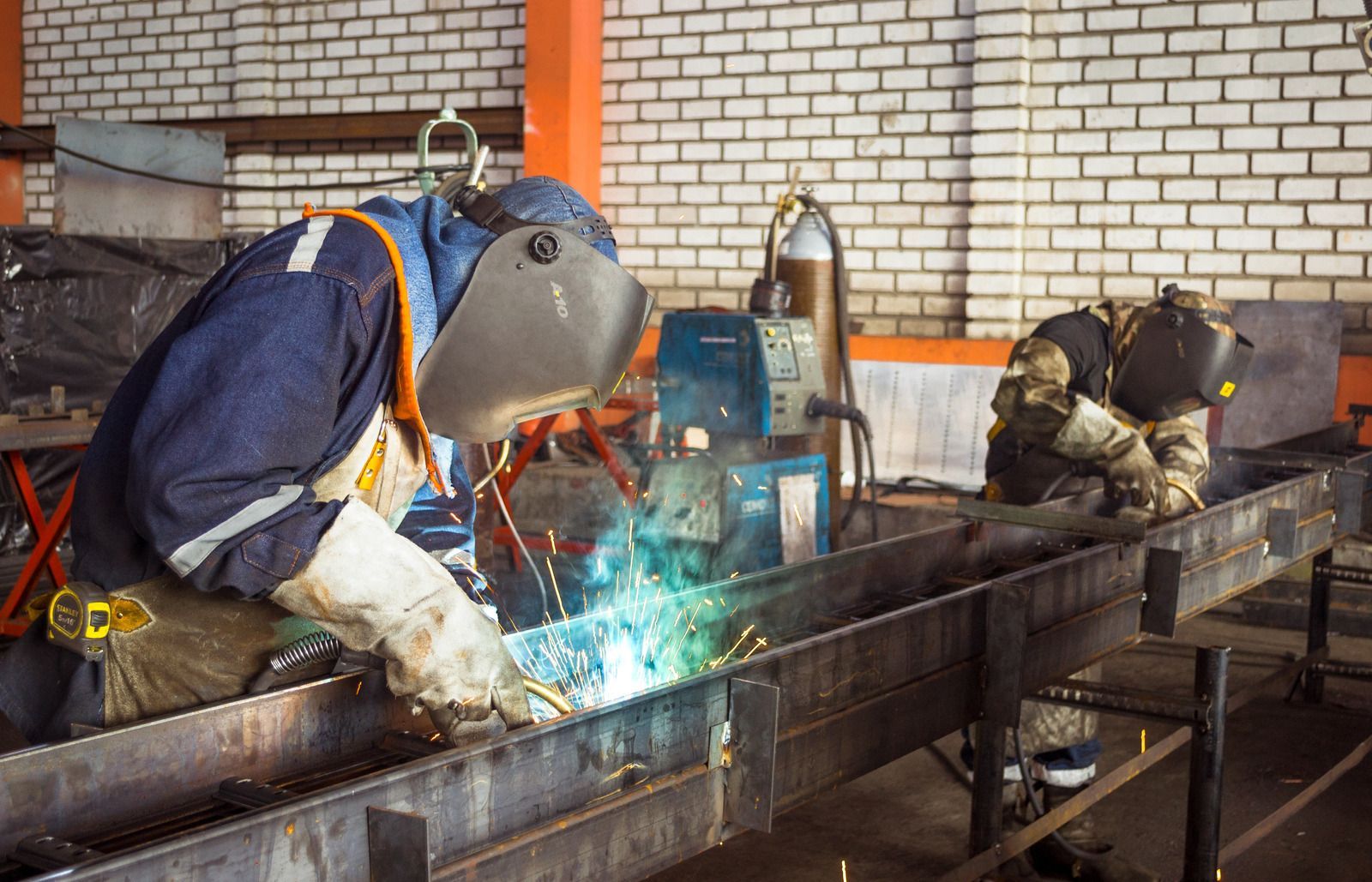Two welders in protective gear welding metal beams in a workshop.
