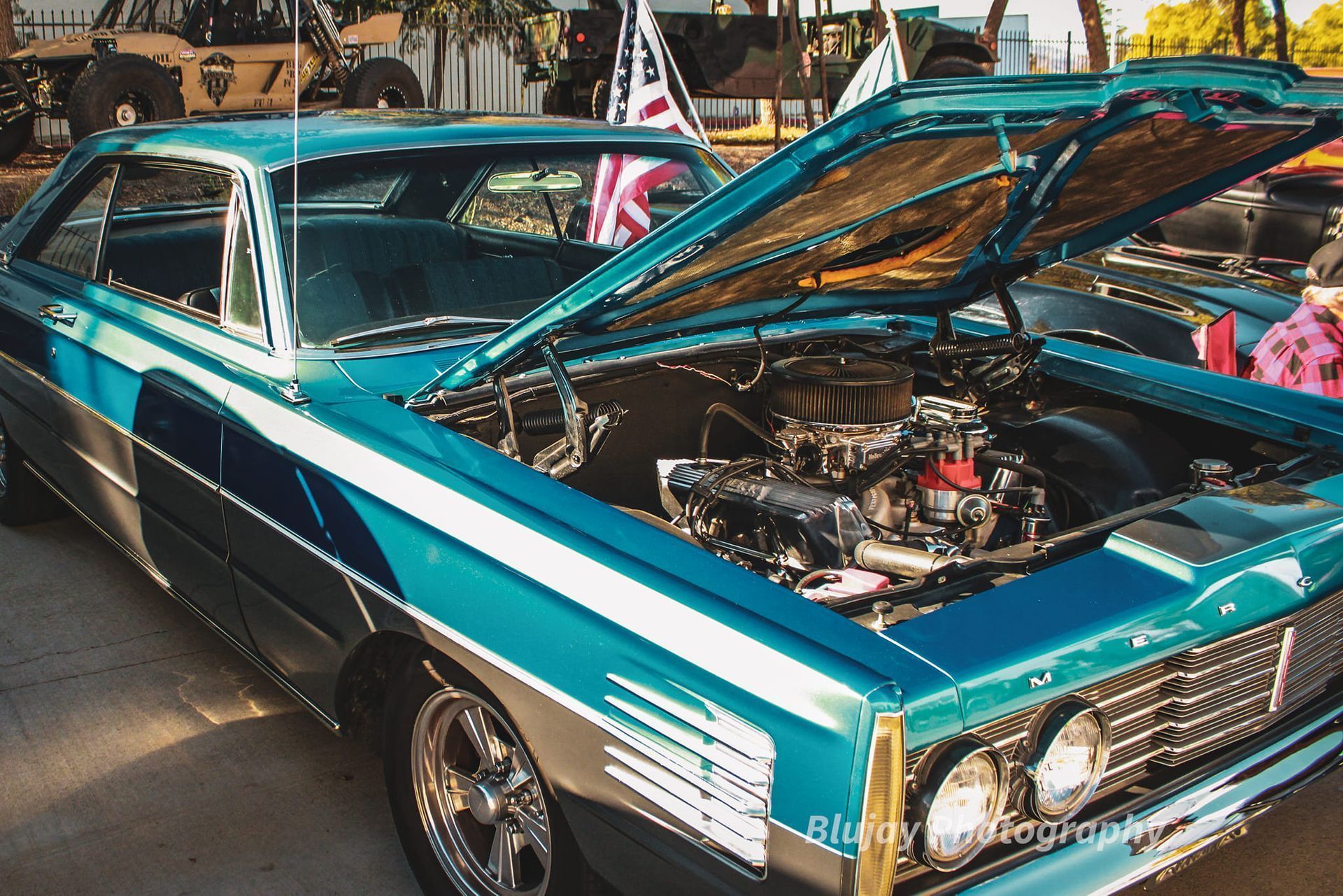 Teal classic car with open hood, engine exposed. Chrome accents, American flag in background.