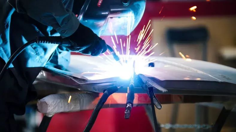 Welder using torch, sparks flying, working on a silver car panel in a workshop.
