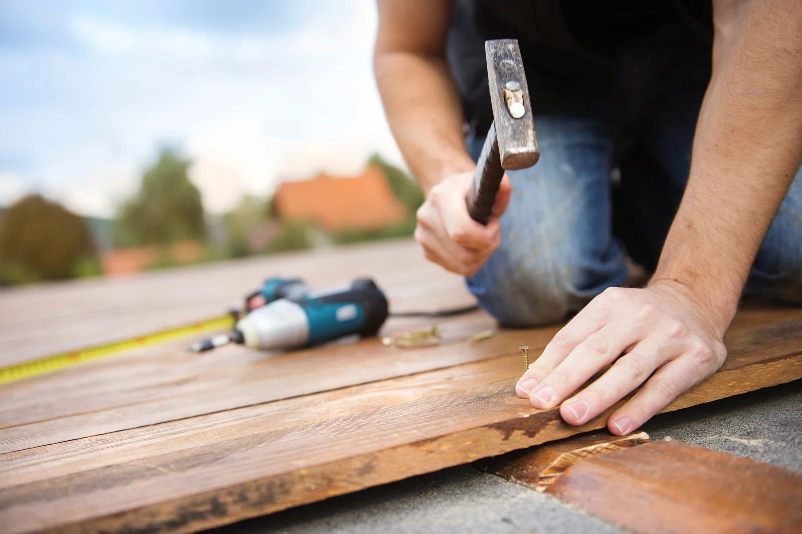 Person kneeling, hammering a nail into wooden planks on an outdoor deck; drill and tape measure visible.