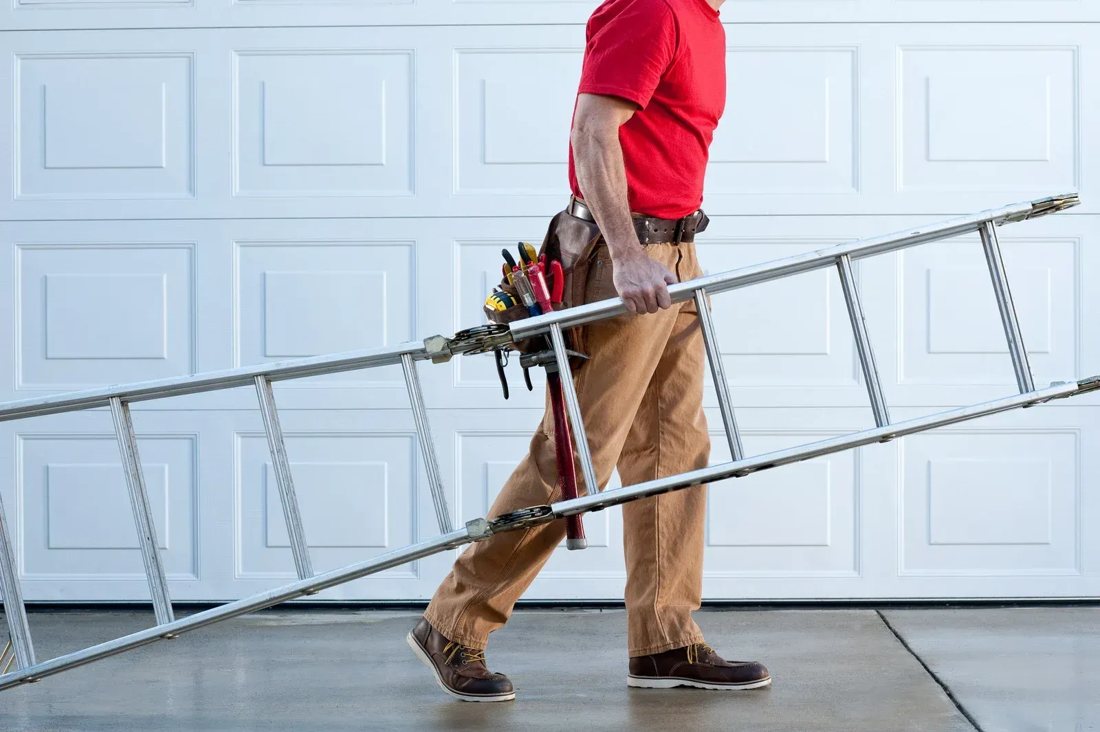 Man carrying a ladder, walking in front of a white garage door. He wears a red shirt and brown pants.