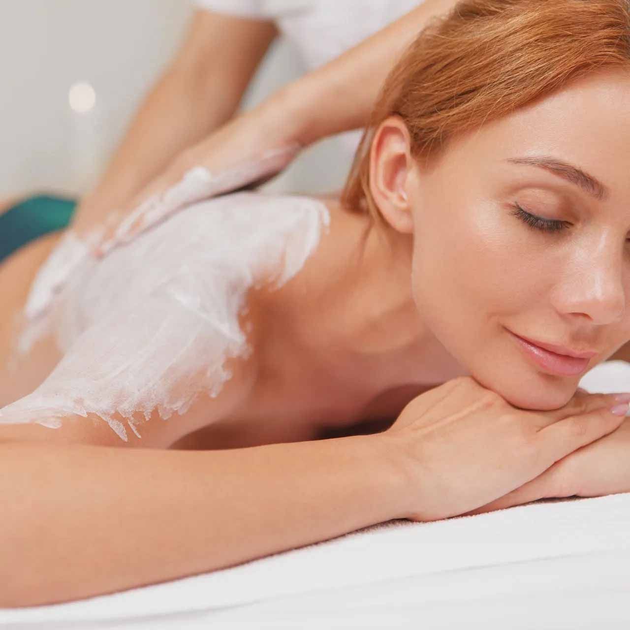 Une femme se fait gommage du corps dans un spa, le dos recouvert de crème, les yeux fermés en signe de détente.