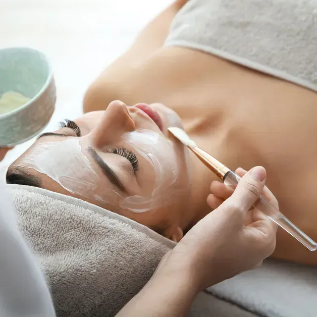 Une femme recevant un soin du visage, avec un pinceau appliquant une crème blanche dans un cadre de spa.
