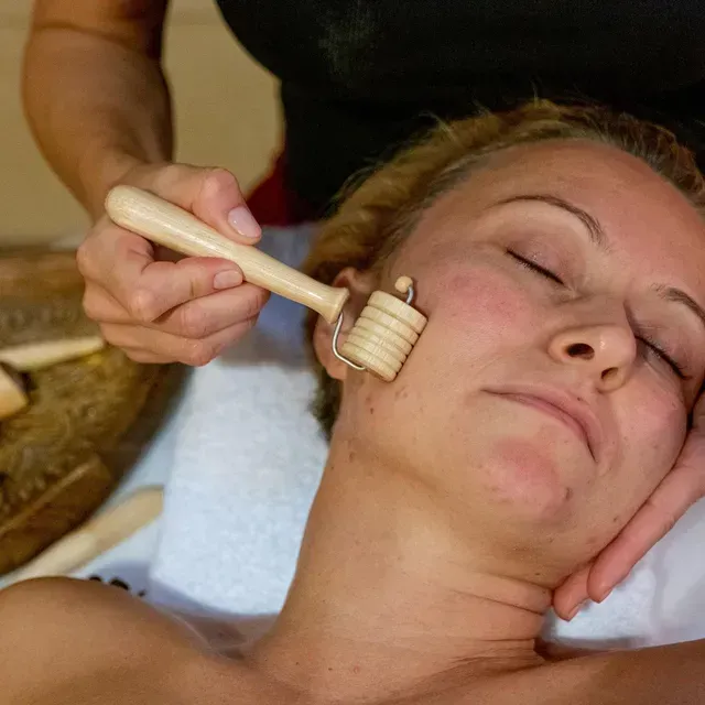 Femme recevant un massage du visage avec un rouleau en bois, les yeux fermés, à l'intérieur.