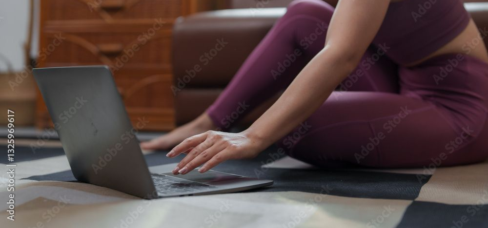 Une femme en tenue de sport violette utilise un ordinateur portable sur un tapis ; cadre domestique.